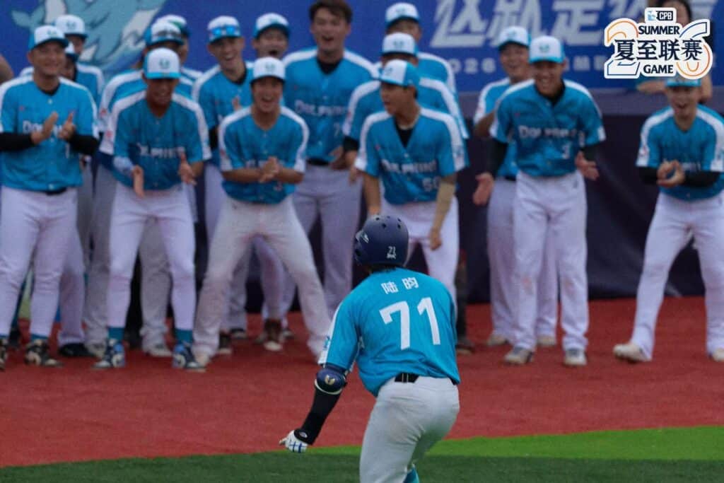 Chinese baseball player in blue uniform on field with team celebrating behind.